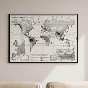 May include: A black and white vintage world map poster with the title "Distribution Annuelle des Pluies" framed in a black frame. The map shows the annual rainfall distribution around the world. The poster is hanging on a white wall above a beige couch.