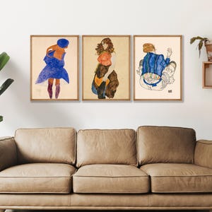 Könnte beinhalten: Drei gerahmte Kunstdrucke in einer Reihe. Der linke Druck zeigt eine Figur in blauem Mantel und dunklen Stiefeln. Der mittlere Druck zeigt eine Figur in braun-orangem Outfit. Der rechte Druck zeigt eine sitzende Figur in einer blau gestreiften Jacke.