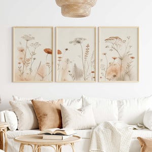 May include: A set of three rectangular wall art prints featuring delicate botanical illustrations of wildflowers and grasses in shades of beige, cream, and muted orange, each enclosed in a light-colored wooden frame and displayed in a row.