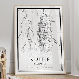 May include: A framed black and white map of Seattle, Washington, with coordinates, sits next to a woven rattan chair and a round jute rug. An open book and a blanket are on the chair.