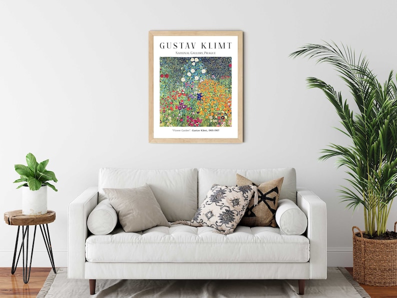 Op de afbeelding: Een ingelijste print van Gustav Klimts schilderij "Bloementuin", met een levendige weergave van kleurrijke bloemen en een donkerblauwe lucht. De print is opgehangen aan een witte muur boven een witte bank met kussens.