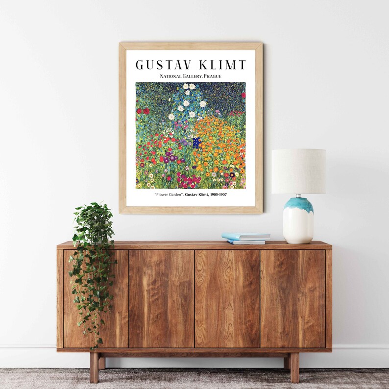 Op de afbeelding: Een ingelijste print van Gustav Klimts schilderij "Bloementuin", met een levendig veld vol kleurrijke bloemen. De print wordt tentoongesteld op een houten plank met een lamp en een plant in een pot.