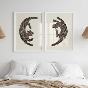 May include: Two black and white stylized illustrations of a tiger in a circular pose, framed in white and hung on a white wall above a bed with white bedding.