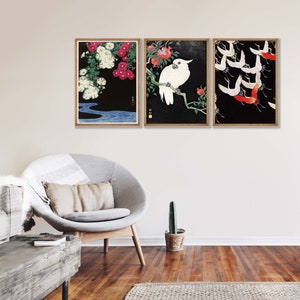 K&ouml;nnte beinhalten: Drei gerahmte Drucke mit japanisch inspirierten Kunstwerken. Der erste Druck zeigt wei&szlig;e Blumen auf schwarzem Hintergrund mit einem blauen Fluss. Der zweite Druck zeigt einen wei&szlig;en Kakadu, der auf einem Ast mit roten Granat&auml;pfeln sitzt. Der dritte Druck zeigt rote und wei&szlig;e Kraniche, die vor einem schwarzen Hintergrund fliegen.
