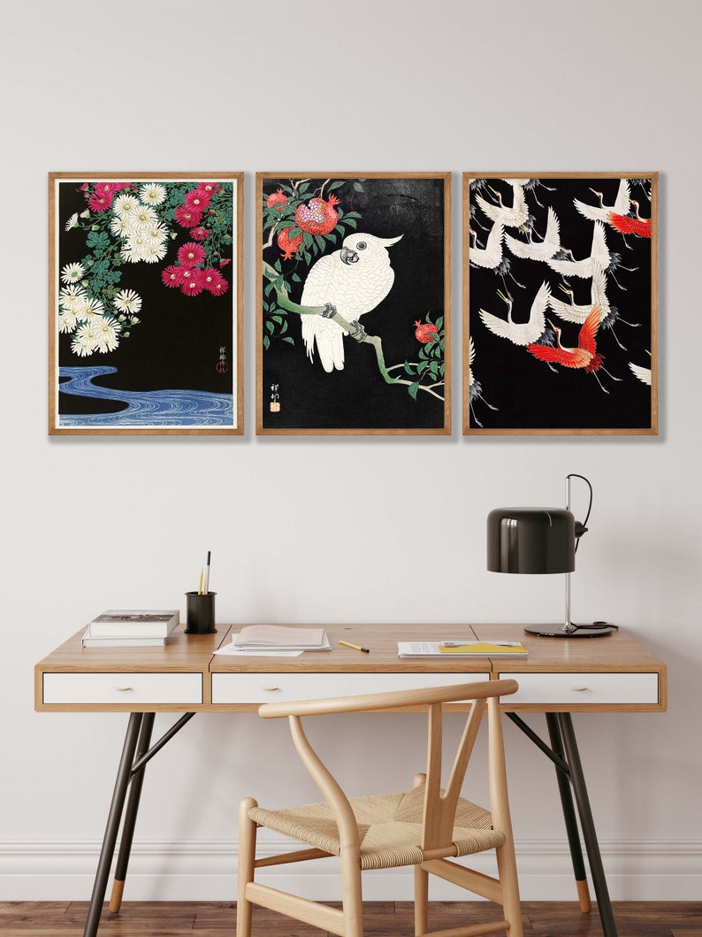 K&ouml;nnte beinhalten: Drei gerahmte Drucke mit japanisch inspirierten Kunstwerken. Der linke Druck zeigt wei&szlig;e und rosa Blumen auf schwarzem Hintergrund mit einem blauen Fluss am unteren Rand. Der mittlere Druck zeigt einen wei&szlig;en Kakadu, der auf einem Ast mit roten Granatapfel Fr&uuml;chten sitzt. Der rechte Druck zeigt eine Schar roter und wei&szlig;er Kraniche, die vor einem schwarzen Hintergrund fliegen.