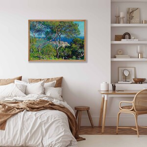 May include: A framed painting of a coastal landscape with a view of a town and the sea from a hillside. The painting features a vibrant blue sky, lush green trees, and a white town with buildings. The painting is framed in a light brown wood frame.