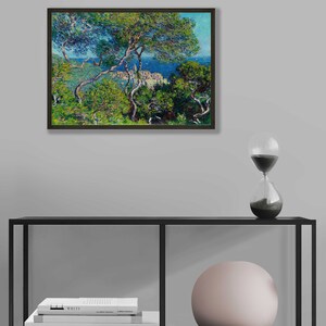 May include: A framed painting of a landscape with a view of a town on a hill overlooking the ocean. The painting is done in a style reminiscent of Impressionism, with visible brushstrokes and a focus on light and color. The painting is framed in a dark wood frame.