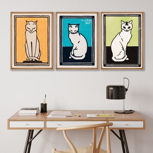 May include: Three framed vintage prints of white cats with black outlines. The cats are sitting with their tails curled around their legs. The frames are brown wood with a white mat. The prints are on a white wall above a wooden desk with a chair and a lamp.