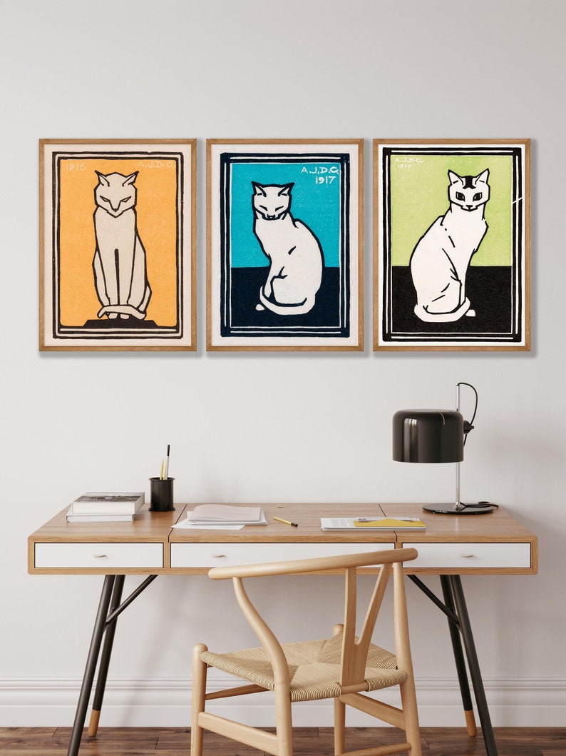May include: Three framed vintage prints of white cats with black outlines. The cats are sitting with their tails curled around their legs. The frames are brown wood with a white mat. The prints are on a white wall above a wooden desk with a chair and a lamp.