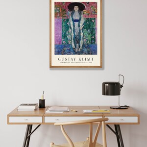 Pu&ograve; includere: Un poster incorniciato di un dipinto di Gustav Klimt intitolato "Ritratto di Adele Bloch-Bauer, 1907". Il dipinto raffigura una donna con un lungo abito fluido con un motivo floreale. Lo sfondo &egrave; un mix vibrante di colori e motivi.