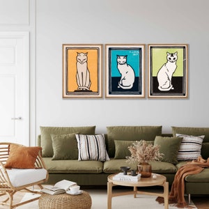 May include: Three framed prints of stylized black and white cat illustrations. The cats are sitting and facing forward. The frames are wood with a light brown finish. The background is a white wall.