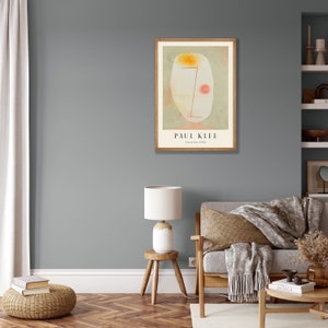 May include: A framed art print featuring a Paul Klee painting titled Untitled, 1934. The artwork displays an abstract, pale oval face with a single red dot, a yellow patch, and thin red lines on a muted green background, all set within a light wood frame.