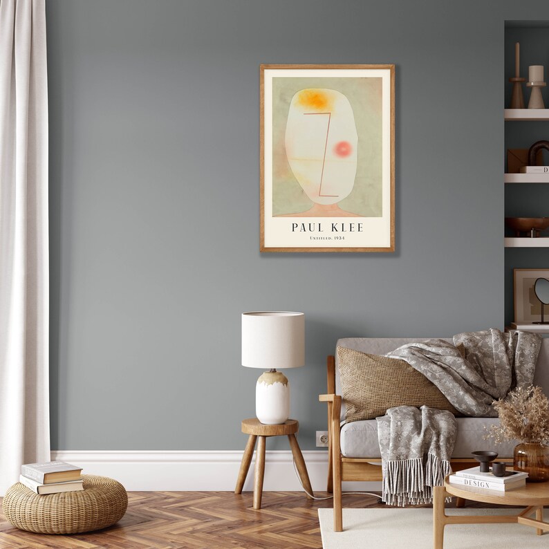 May include: A framed art print featuring a Paul Klee painting titled Untitled, 1934. The artwork displays an abstract, pale oval face with a single red dot, a yellow patch, and thin red lines on a muted green background, all set within a light wood frame.