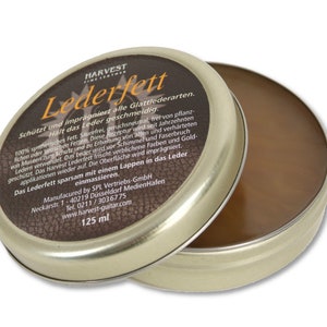 May include: A round tin container of Harvest Leather Grease, a product for protecting and conditioning leather. The tin is gold with a brown label that reads "Harvest Lederfett" and includes instructions for use. The tin contains a brown, thick, leather grease.