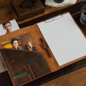 Può includere: Una cartella in pelle marrone con un blocco note, una penna e tasche. La cartella è aperta, rivelando un blocco note bianco con una clip argentata. Una penna poggia sul blocco note. Riviste e carte sono nelle tasche.