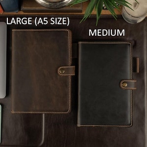 May include: Two leather journals, one large and one medium, are displayed on a brown surface. The large journal is brown with a button closure and the text "LARGE (A5 SIZE)". The medium journal is black with a button closure and the text "MEDIUM".