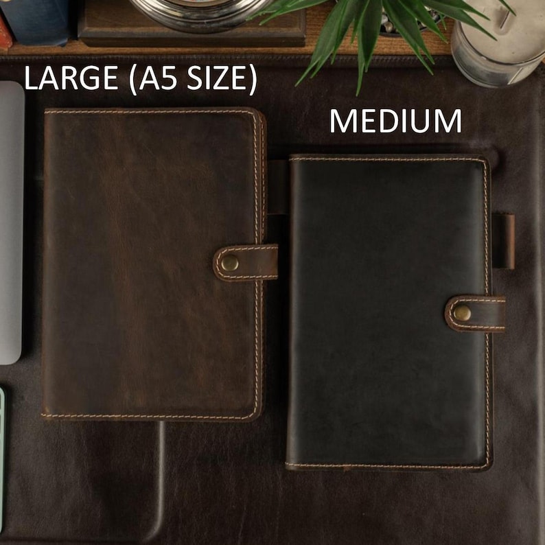 May include: Two leather journals, one large and one medium, are displayed on a brown surface. The large journal is brown with a button closure and the text "LARGE (A5 SIZE)". The medium journal is black with a button closure and the text "MEDIUM".