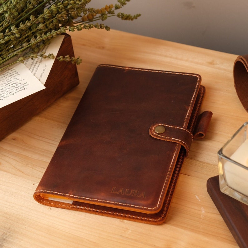A brown leather bound journal with a flap closure and a brass button. The journal has a stitched edge and the name 'LAURA' is embossed on the front cover for sample.