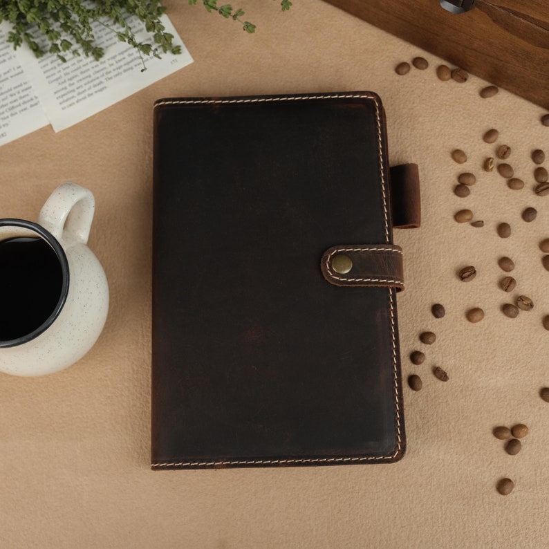 A dark brown leather bound journal with a flap closure and a stitched design. The journal has a brown leather strap with a brass button closure.