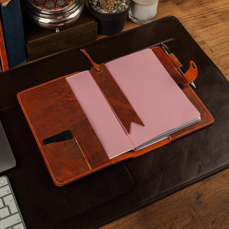 May include: Open leather journal with a pink notebook inside, a brown bookmark, and a pen. The journal is open on a brown leather desk mat. The journal and bookmark are a similar shade of brown.