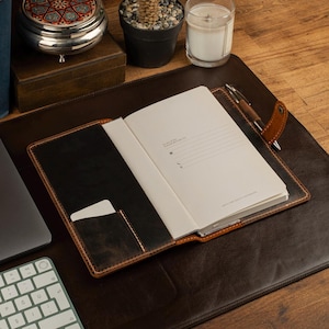 May include: Open leather journal with a pen, resting on a dark brown leather desk mat. The journal has a black interior with a card pocket. A white keyboard and laptop are visible in the background. A small potted plant, candle, and decorative box are also on the desk.