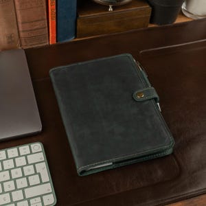 May include: A dark green leather journal with a pen secured by a loop. The journal is closed with a snap closure. A laptop and keyboard are visible in the background, suggesting a workspace.