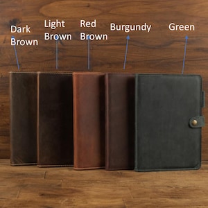 May include: A selection of leather journals in shades of dark brown, light brown, red brown, burgundy, and green. Each journal is rectangular with stitched edges. The green journal has a button clasp.