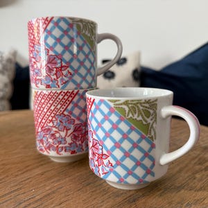 May include: Three white ceramic mugs with colorful patchwork designs. The mugs feature patterns of red floral, blue plaid, and green floral. One mug is stacked on top of two others. The mugs have white handles and are sitting on a wooden surface.