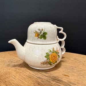 May include: A cream-colored ceramic tea-for-one set with a speckled finish. The teapot has a spout and a handle, and the cup sits on top. Both pieces are decorated with yellow and green floral designs.