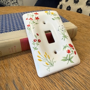 May include: White ceramic light switch cover with a floral design featuring red, yellow, and green flowers and foliage. The cover has a rectangular opening for the switch and two screw holes.