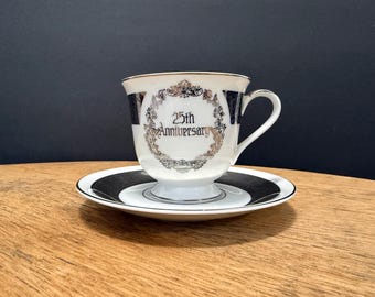 May include: A white teacup and saucer set with a gold rim. The teacup features the text "25th Anniversary" in a decorative gold frame. The saucer has a black and white concentric circle design.