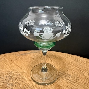 May include: Clear glass candle holder with a green base and stem. The bowl of the holder is decorated with white shamrocks and a Claddagh design. The candle holder sits on a wooden surface.
