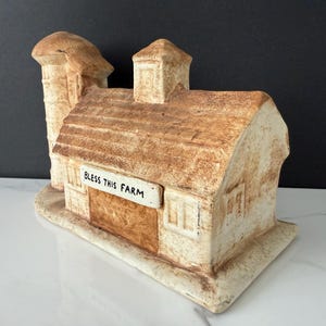 May include: A ceramic farm figurine in shades of beige and brown, featuring a barn with a silo. The words "BLESS THIS FARM" are displayed on a small sign. The figurine has a rustic, aged appearance.