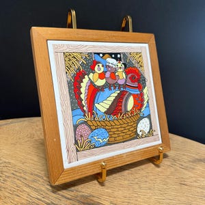 May include: A framed ceramic tile featuring a colorful illustration of chickens in a basket with eggs. The artwork is set in a light brown wooden frame and displayed on a gold-colored stand.