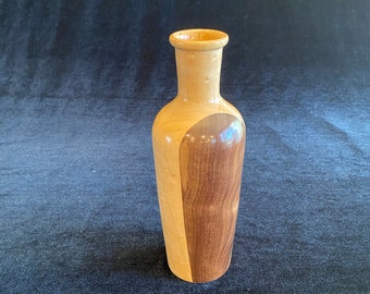 May include: A wooden vase with a light brown and dark brown wood grain pattern. The vase is tall and slender with a rounded bottom and a slightly flared opening.
