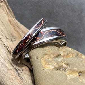 May include: Two silver rings with a dark, iridescent inlay and copper-colored wire detailing. The rings are set against a natural background of wood and stone, showcasing their intricate design and craftsmanship. The rings are likely wedding bands or fashion accessories.
