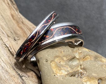May include: Two silver rings with a dark, iridescent inlay and copper-coloured wire detailing. The rings are set against a natural background of wood and stone, showcasing their intricate design and craftsmanship. The rings are likely wedding bands or fashion accessories.