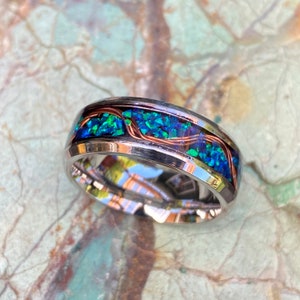 An 8mm wide stainless steel ring rests on a green and brown stone background.  The ring has blue opal inlayed with a strand of copper winding in a sine wave pattern through the channel.