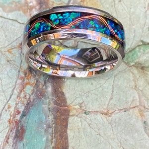 An 8mm wide stainless steel ring rests on a green and brown stone background.  The ring has blue opal inlayed with a strand of copper winding in a sine wave pattern through the channel.