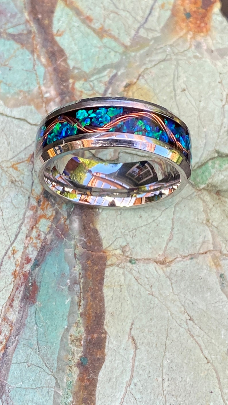 An 8mm wide stainless steel ring rests on a green and brown stone background.  The ring has blue opal inlayed with a strand of copper winding in a sine wave pattern through the channel.