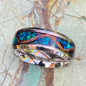 An 8mm wide stainless steel ring rests on a green and brown stone background.  The ring has blue opal inlayed with a strand of copper winding in a sine wave pattern through the channel.