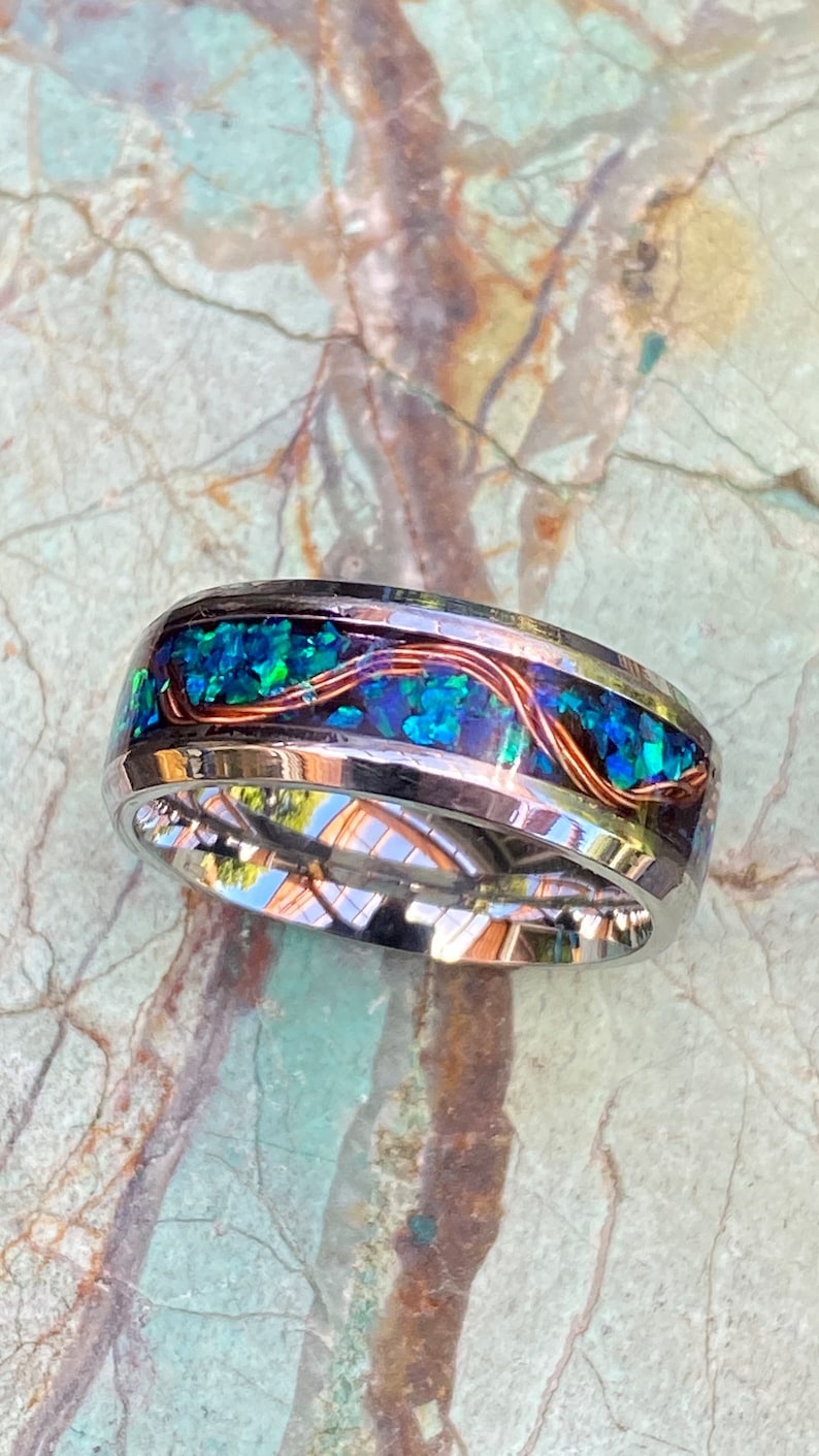 An 8mm wide stainless steel ring rests on a green and brown stone background.  The ring has blue opal inlayed with a strand of copper winding in a sine wave pattern through the channel.