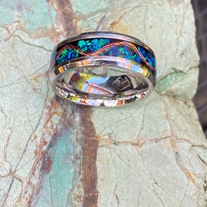 An 8mm wide stainless steel ring rests on a green and brown stone background.  The ring has blue opal inlayed with a strand of copper winding in a sine wave pattern through the channel.