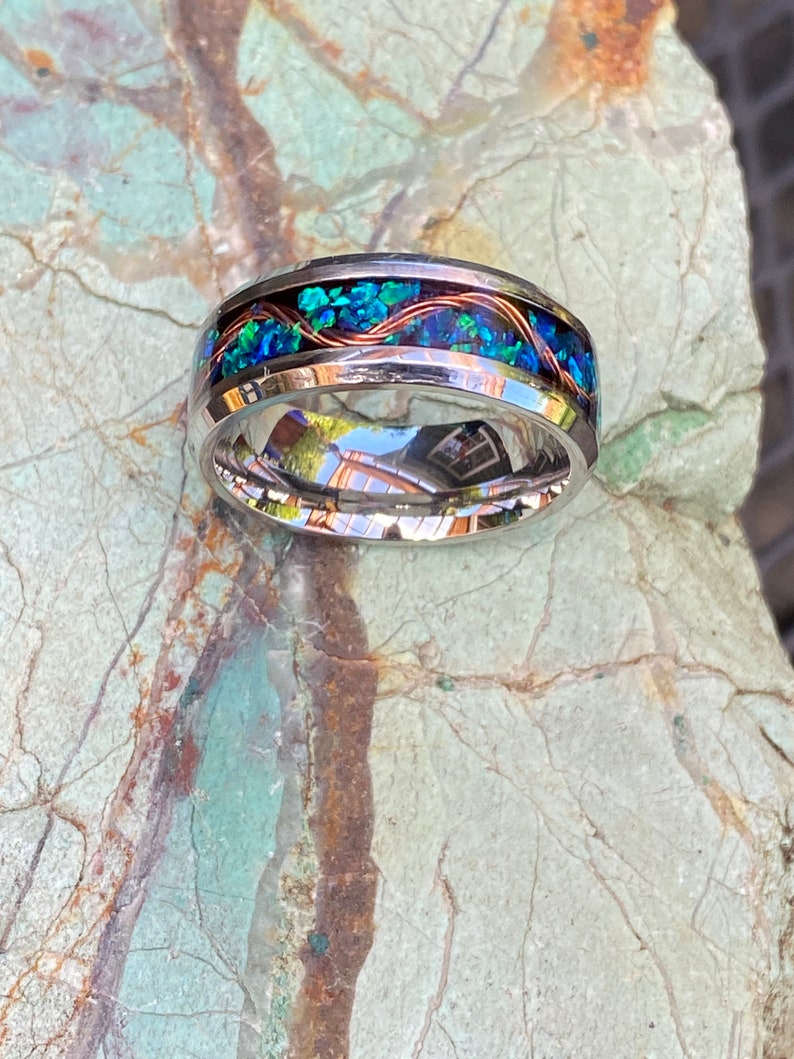 An 8mm wide stainless steel ring rests on a green and brown stone background.  The ring has blue opal inlayed with a strand of copper winding in a sine wave pattern through the channel.