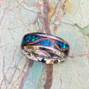 An 8mm wide stainless steel ring rests on a green and brown stone background.  The ring has blue opal inlayed with a strand of copper winding in a sine wave pattern through the channel.