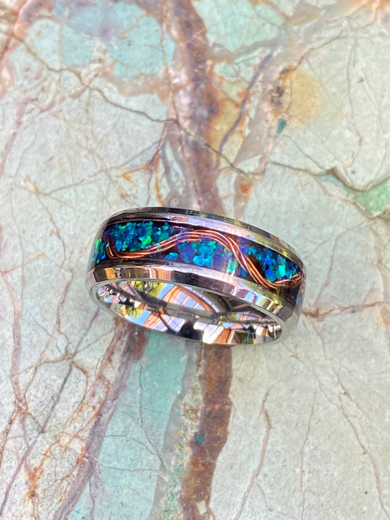 An 8mm wide stainless steel ring rests on a green and brown stone background.  The ring has blue opal inlayed with a strand of copper winding in a sine wave pattern through the channel.
