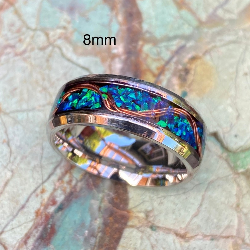 An 8mm wide stainless steel ring rests on a green and brown stone background.  The ring has blue opal inlayed with a strand of copper winding in a sine wave pattern through the channel.