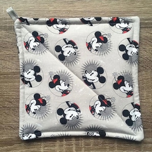 May include: Square gray pot holder featuring a repeating pattern of Mickey Mouse in a circular design with a sunburst background. The pot holder has a loop for hanging and a white border.