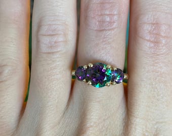 May include: A gold ring with three oval gemstones that change colour. The gemstones display purple and green tones. The ring is set on a gold band, highlighting the stones' vibrant colours.
