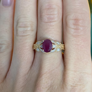 May include: A gold ring with a large oval ruby gemstone at the centre, encircled by smaller white gemstones. The band has an intricate, ornate design with leaf-like patterns. The ring is on a person's finger.